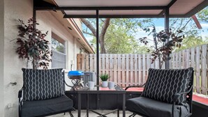 Eine überdachte Terrasse mit zwei schwarzen Stühlen mit gemusterten Kissen, einem Tisch mit einem blauen Grill und Blumentöpfen.