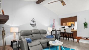 Ein modernes Wohnzimmer mit einem grauen Sofa, einem kleinen runden Tisch und einem durch eine offene Tür einsehbaren Küchenbereich.