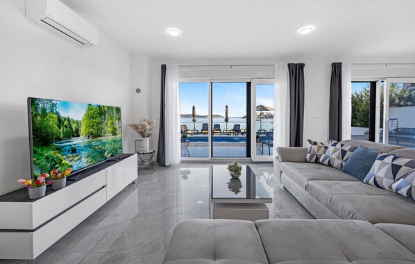 Un salon moderne doté d’un téléviseur à écran plat, d’un canapé, d’une table basse et offrant une vue sur une piscine et un espace de détente extérieur.