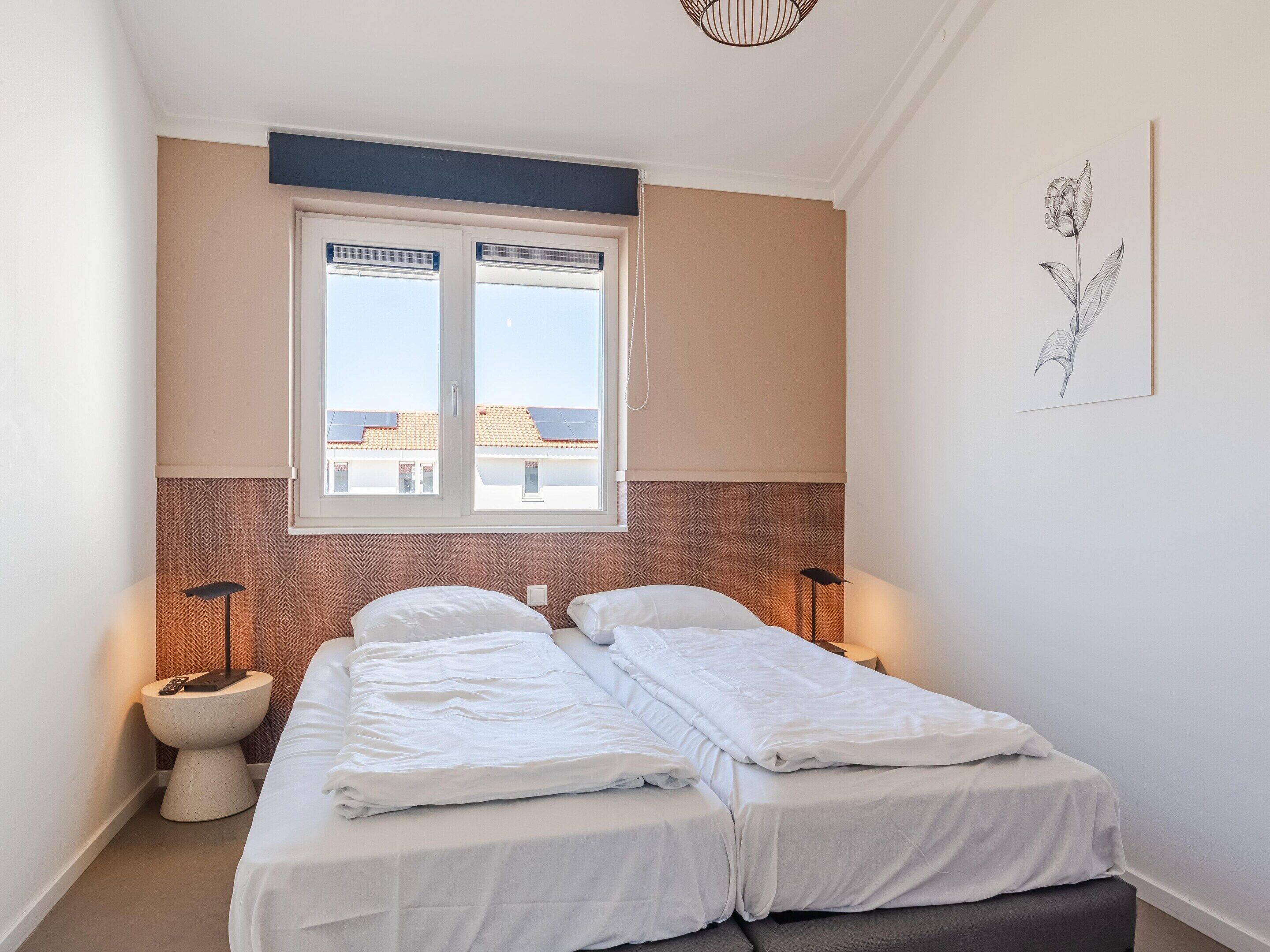 A bedroom with two beds, a window with blinds, a bedside table with a lamp, and a wall-mounted artwork.