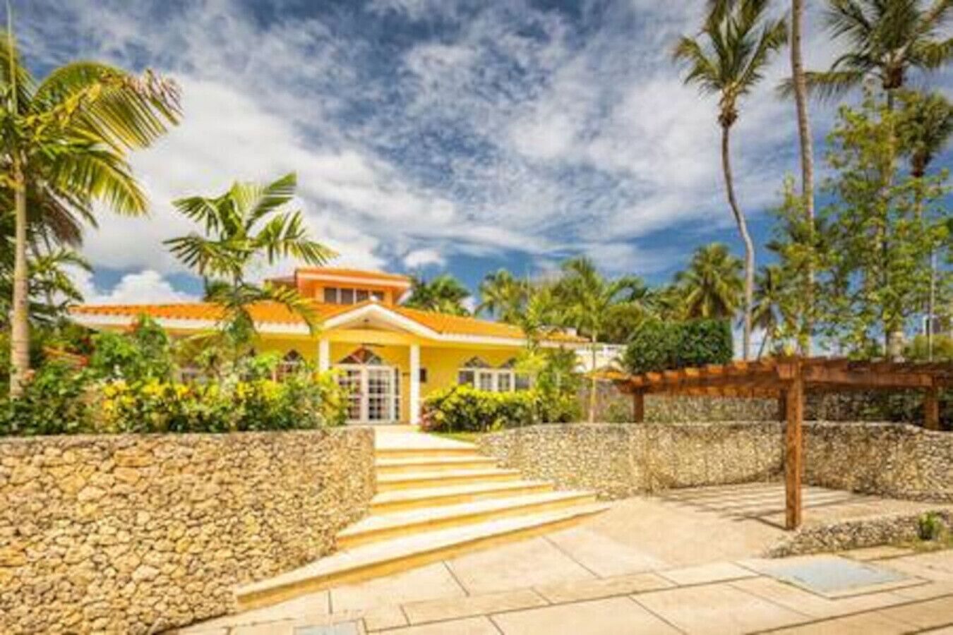 Una casa tropical con pared de piedra, una pérgola de madera y una escalera que lleva a la entrada.