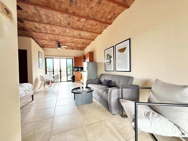 A spacious living area with a grey sofa, a round coffee table, and a dining table with chairs. There is a kitchen in the background, and the room has a high ceiling with exposed brick.
