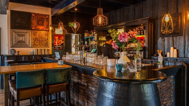 A bar area with a counter, stools, and hanging lights. There are decorative elements on the wall and a person working behind the bar. - Nice Chalet on a Holiday Park 25 km From Amsterdam (Velsen-Zuid)
