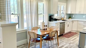 A modern kitchen with white cabinets, a wooden dining table with chairs, and a herringbone-patterned floor. - Historic Atascadero Colony Charmer! (Atascadero)