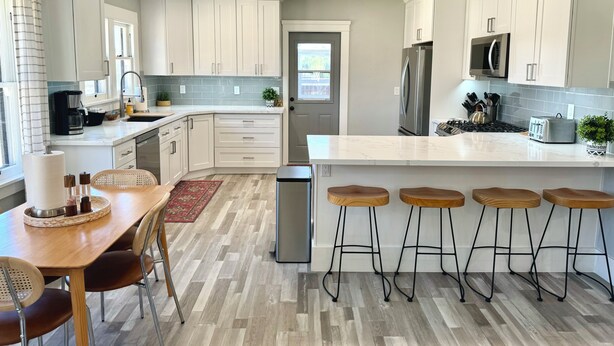 A modern kitchen with white cabinets, a stainless steel refrigerator, a microwave, and a toaster. There are wooden stools and a dining table with chairs. - Historic Atascadero Colony Charmer! (Atascadero)