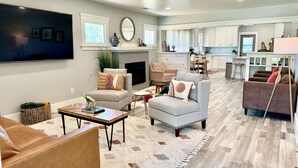 A spacious living room with a fireplace, a television, and a kitchen in the background. - Historic Atascadero Colony Charmer! (Atascadero)
