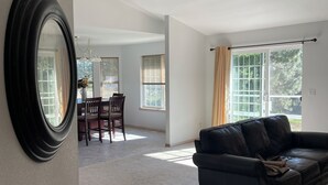 Un salón amplio con un sofá de cuero negro, una zona de comedor con mesa y sillas, y ventanas grandes con persianas.