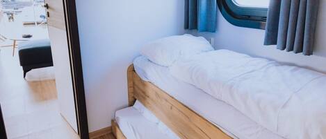 Una cama pequeña y bien hecha con ropa blanca en una habitación de paredes azul claro y ventana con cortinas.