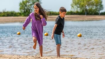 Two children running along a sandy beach with water and buoys in the background.