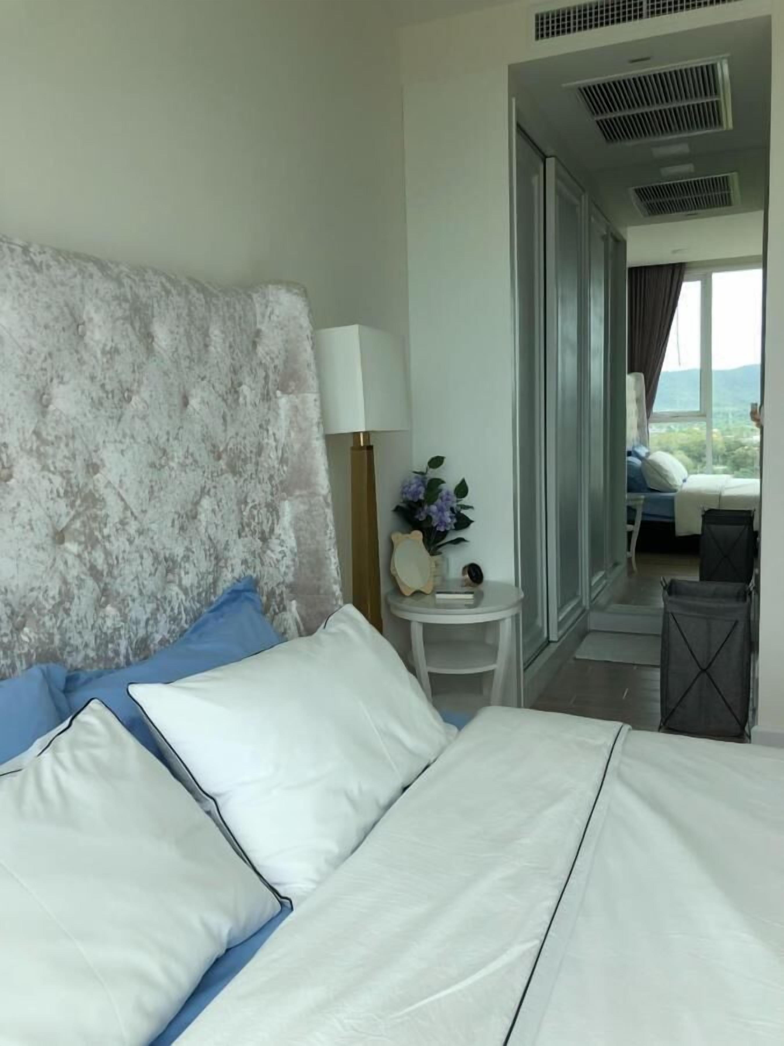 A hotel room with a large bed, a bedside table with a lamp, a small table with a vase of flowers, and a view of another room through an open door.