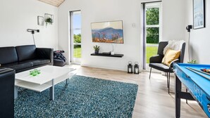 Una sala de estar moderna con un sofá de cuero negro, una mesa de centro blanca, una mesa de billar azul y un ventanal con vistas a zonas verdes.
