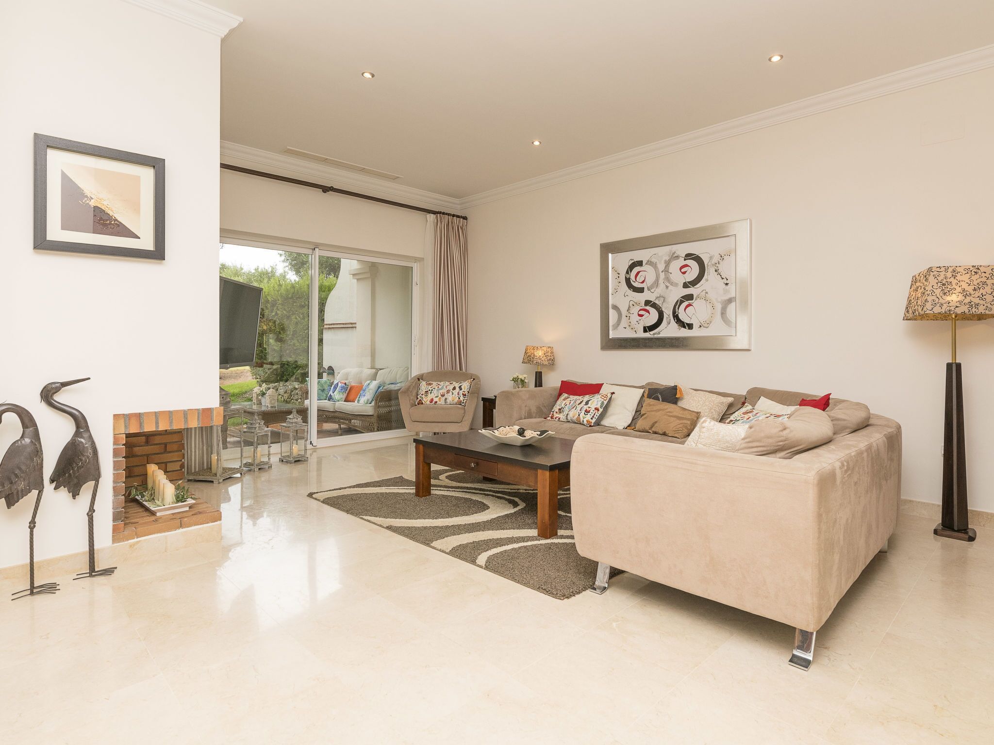 A spacious living room with a beige sofa, a coffee table, and a fireplace. There are two framed artworks on the walls and a floor lamp.