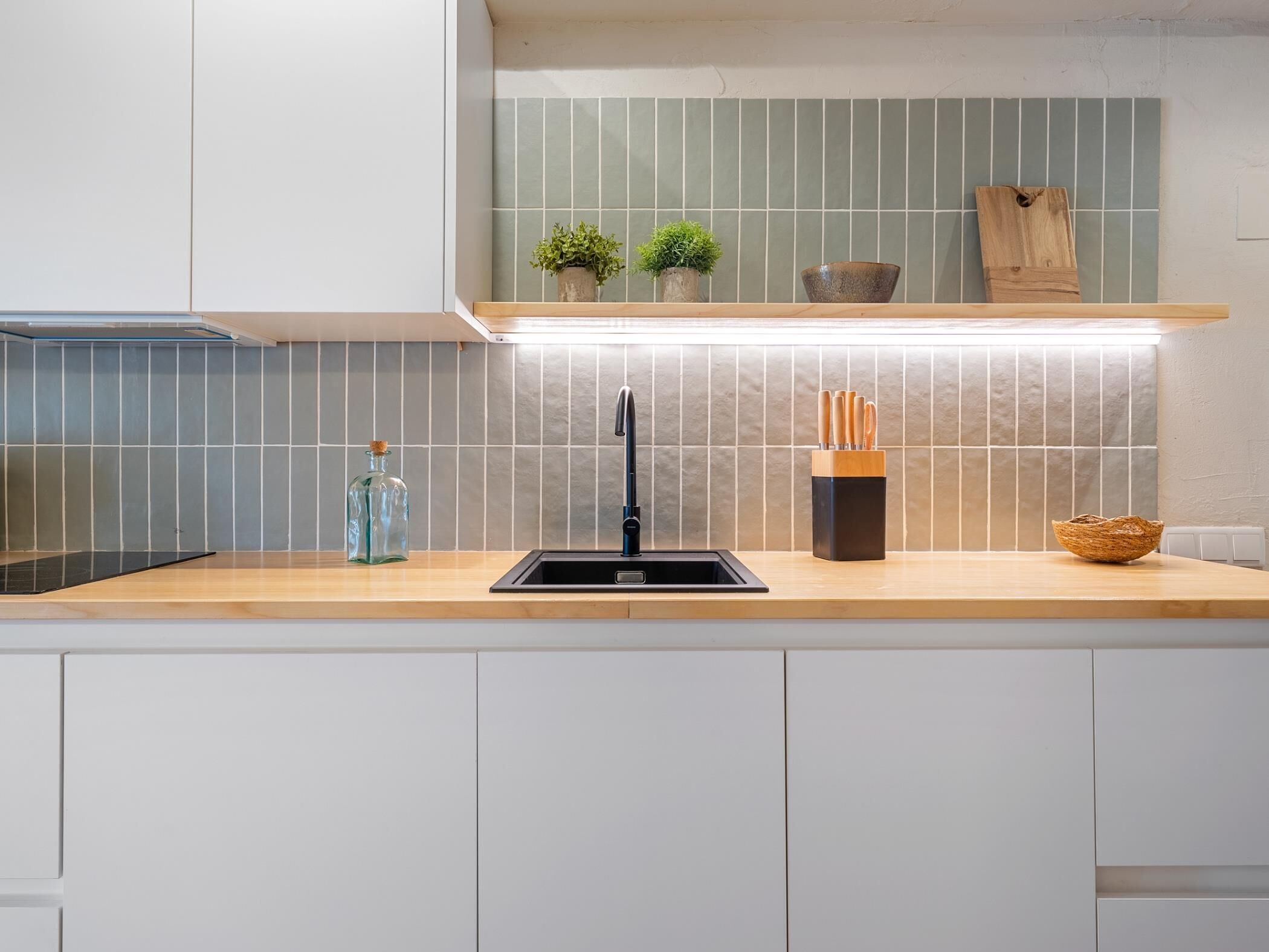A modern kitchen with a black sink, wooden countertop, and a backsplash with green tiles. There are two potted plants on the shelf above the sink and a knife block on the countertop.