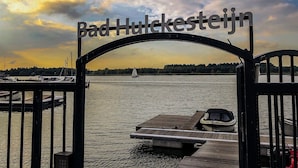 A view through a metal gate with the text "Bad Hulkeestijn" above a body of water, boats docked, and a sailboat in the distance. - Villa With Roof Terrace Near Veluwemeer (Nijkerk)