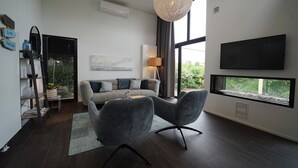 A modern living room with a grey sofa, two armchairs, a large window, a flat-screen TV, and a hanging light fixture. - Villa With Roof Terrace Near Veluwemeer (Nijkerk)