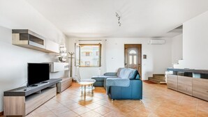A modern living room with a flat-screen TV, a blue sofa, a small round table, and a kitchen area with a wooden cabinet. - GuestHost - Sweet Home in Portoscuso! (Portoscuso)