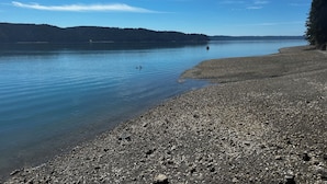 Een kiezelstrand met rustig water en veraf gelegen beboste heuvels.