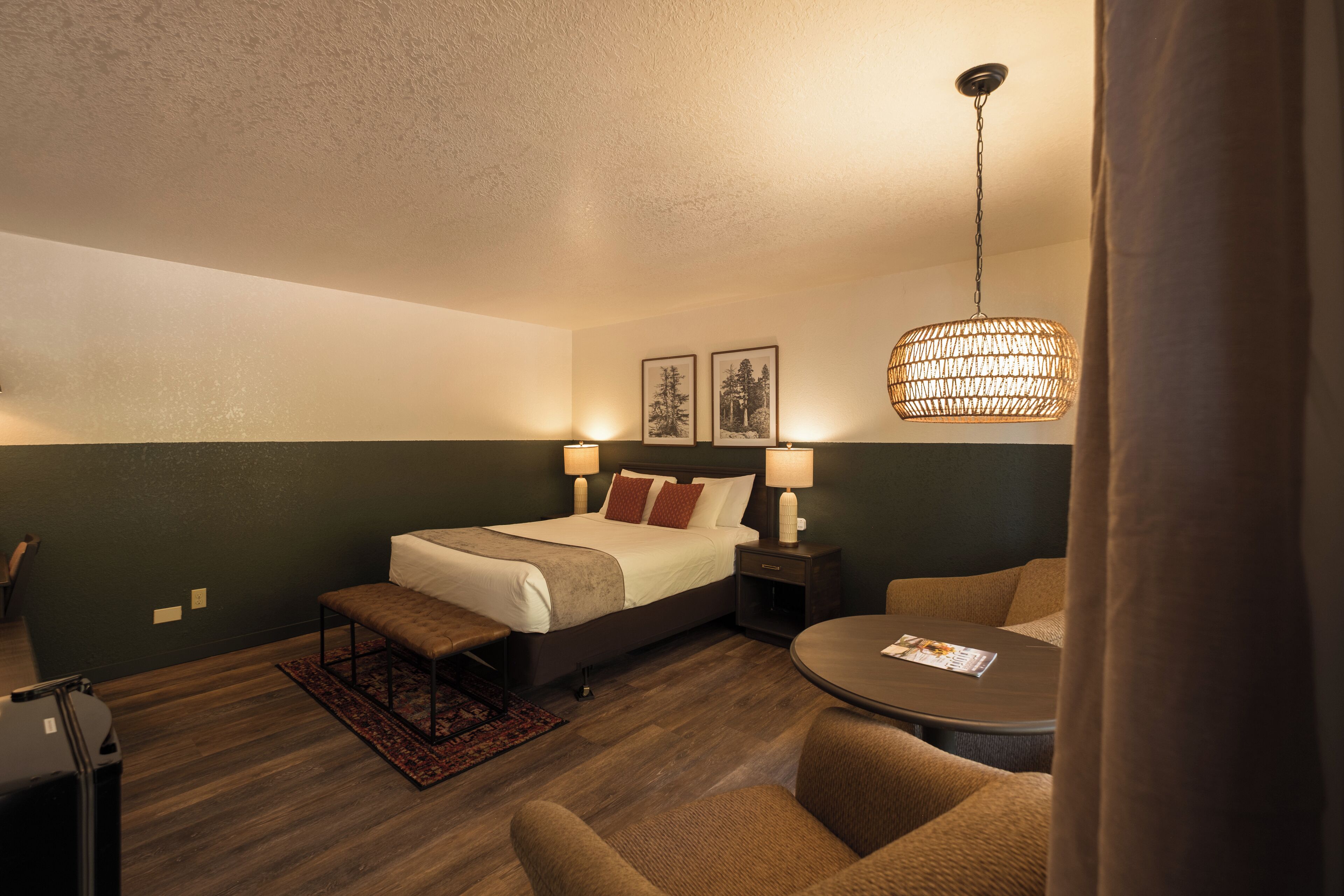 Una habitación de hotel con una cama, mesitas de noche con lámparas, una mesa redonda y una lámpara colgante.