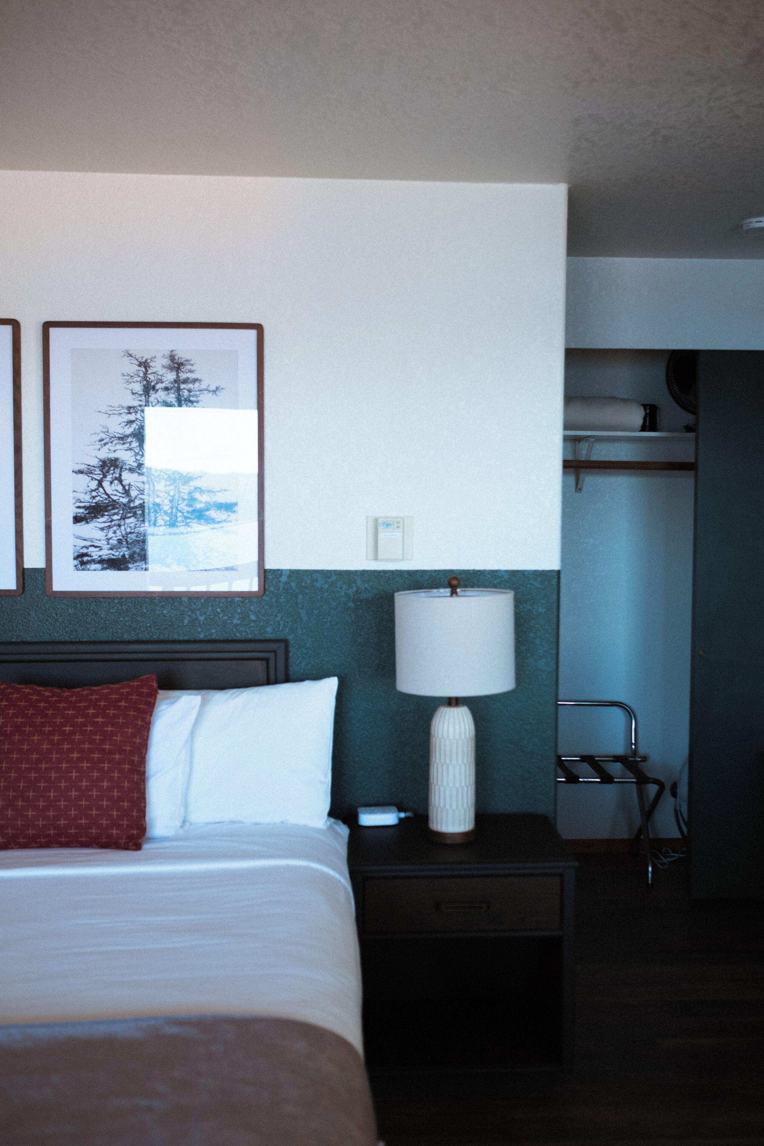 A hotel room with a bed, a nightstand with a lamp, a framed picture on the wall, and a bathroom area with a towel rack.