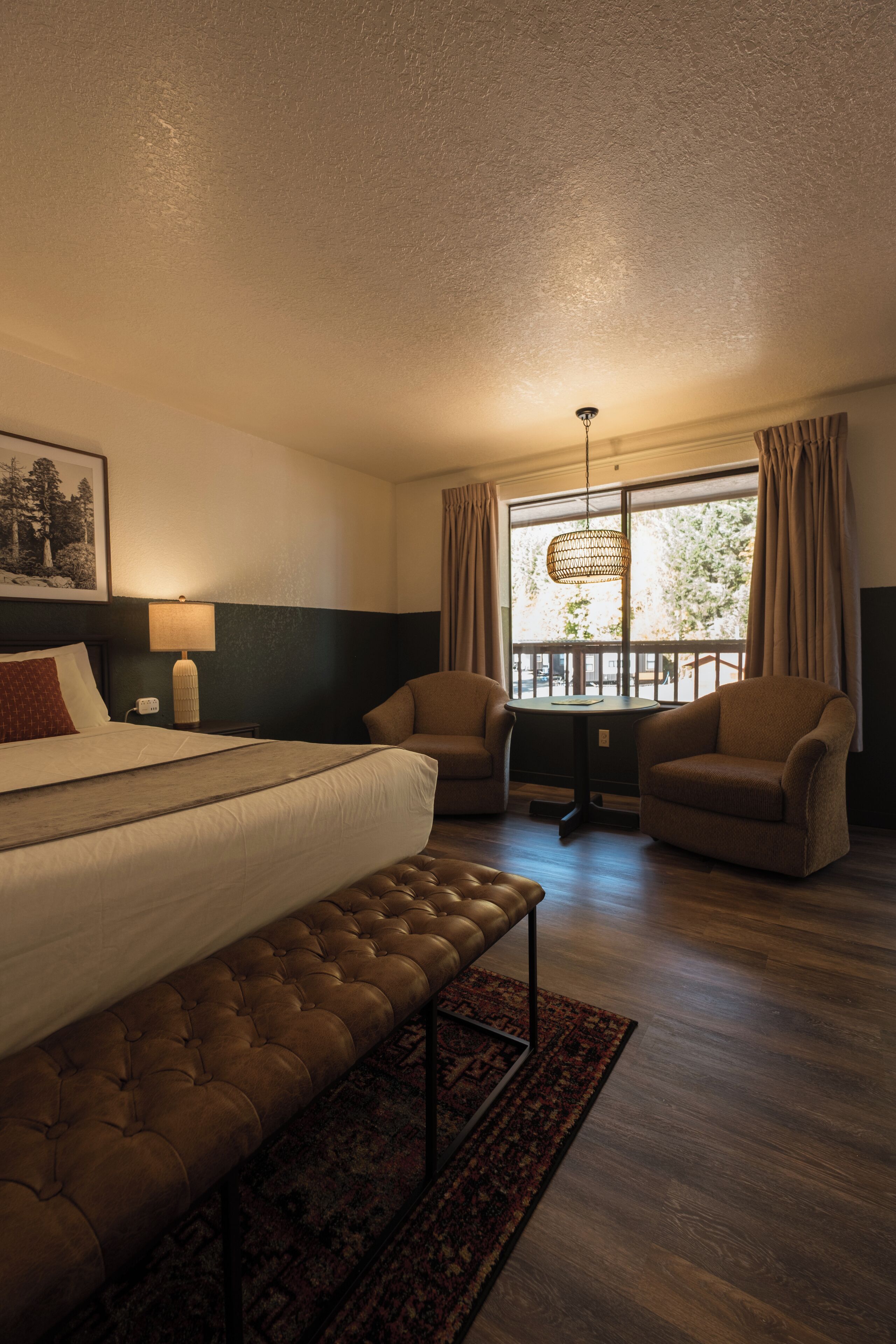 A hotel room with a large bed, two armchairs, a small table, and a view of the outdoors through a window.