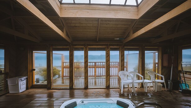 Een houten terras met een jacuzzi, witte stoelen en grote ramen met uitzicht op een wateroppervlak.
