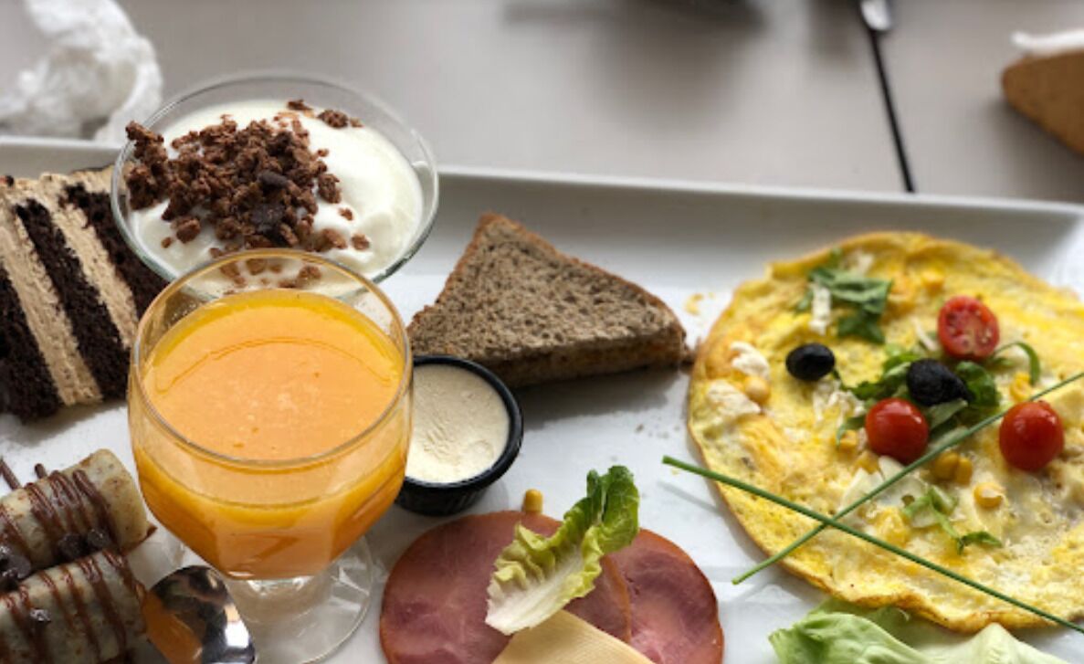 A breakfast plate with a variety of foods including toast, a slice of cake, a glass of orange juice, a bowl of yogurt with chocolate shavings, a small container of cream, a slice of ham, and an omelette with vegetables.