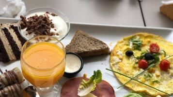A breakfast plate with a variety of foods including toast, a slice of cake, a glass of orange juice, a bowl of yogurt with chocolate shavings, a small container of cream, a slice of ham, and an omelette with vegetables.