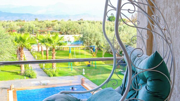 A hanging chair with a cushion, overlooking a pool and a landscaped garden with palm trees and a view of distant mountains. - Jnane Riad (Ghmate)