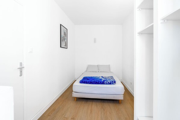 Una habitación estrecha con paredes blancas, una cama, un cuadro en la pared y suelo de madera.