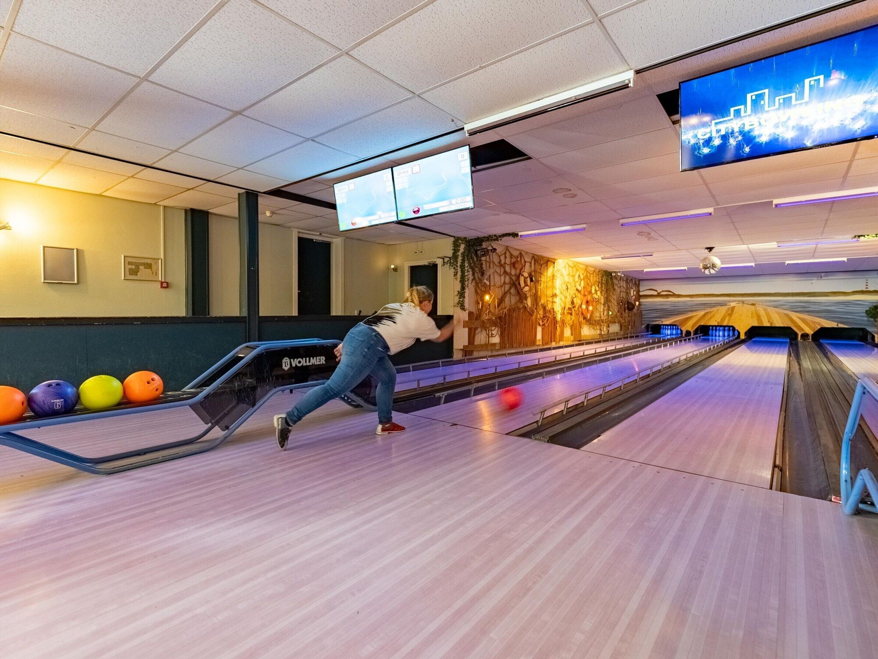 A bowling alley with multiple lanes, a woman bowling, and a large screen displaying a cityscape.