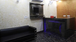 A reception area with a black sofa, a reception desk with blue lighting, an air conditioning unit, and a mounted television. - Hotel Kanak Palace By GRB (New Delhi)