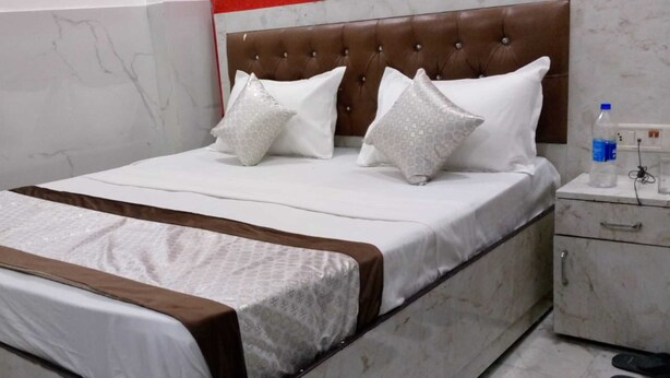 A neatly made bed with a brown headboard, white bedding, and decorative pillows. There is a nightstand with a water bottle and a glass beside the bed. - Hotel Kanak Palace By GRB (New Delhi)