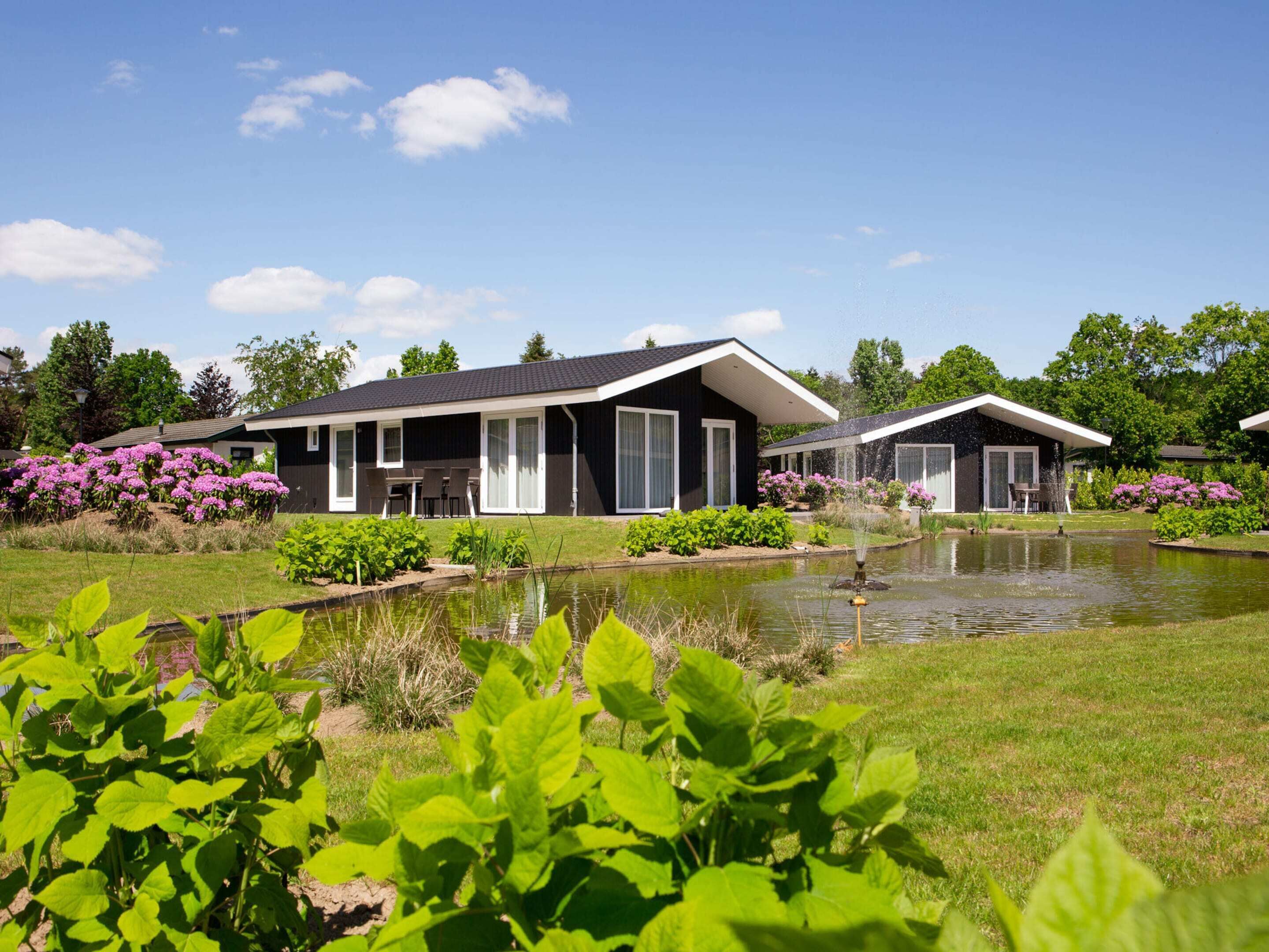 Uma casa moderna de dois andares com um grande gramado frontal, um lago e vegetação ao redor.