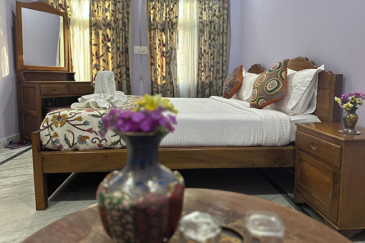 A bedroom with a wooden bed, floral curtains, a wooden nightstand, and a vase with flowers.
