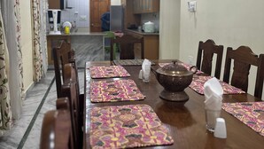 A dining area with a long wooden table set for four, surrounded by chairs. The room has a kitchen in the background with cabinets and a refrigerator. - Jan Mahal (Srinagar)