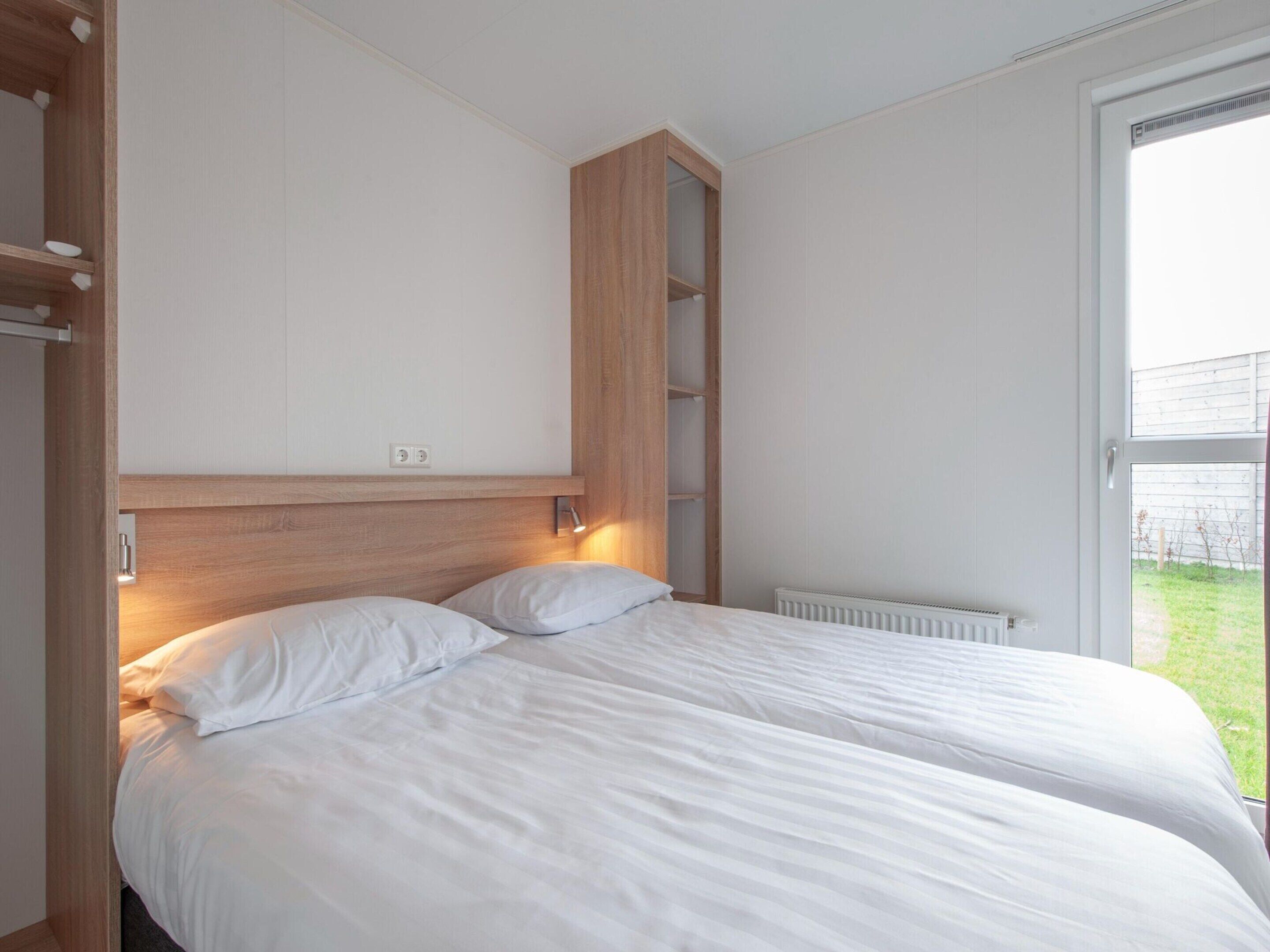 A bedroom with a wooden headboard, two white beds, a built-in shelf, and a view of a garden through a large window.