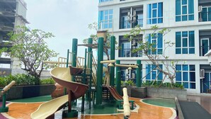 A playground with slides and climbing structures, surrounded by a building with multiple balconies and windows. - Roseville SOHO and Suites by Nagisa Bali (South Tangerang)