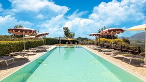 A rectangular swimming pool with lounge chairs and umbrellas, surrounded by a wooden fence and hedges, with a clear sky and distant mountains in the background. - Country House in Marche with Pool & Views (Cagli)