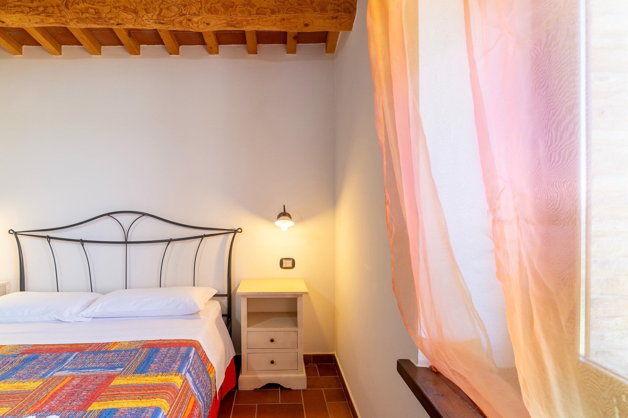 Une chambre à coucher avec un lit, une table de chevet, une lampe et une fenêtre avec des rideaux légers.