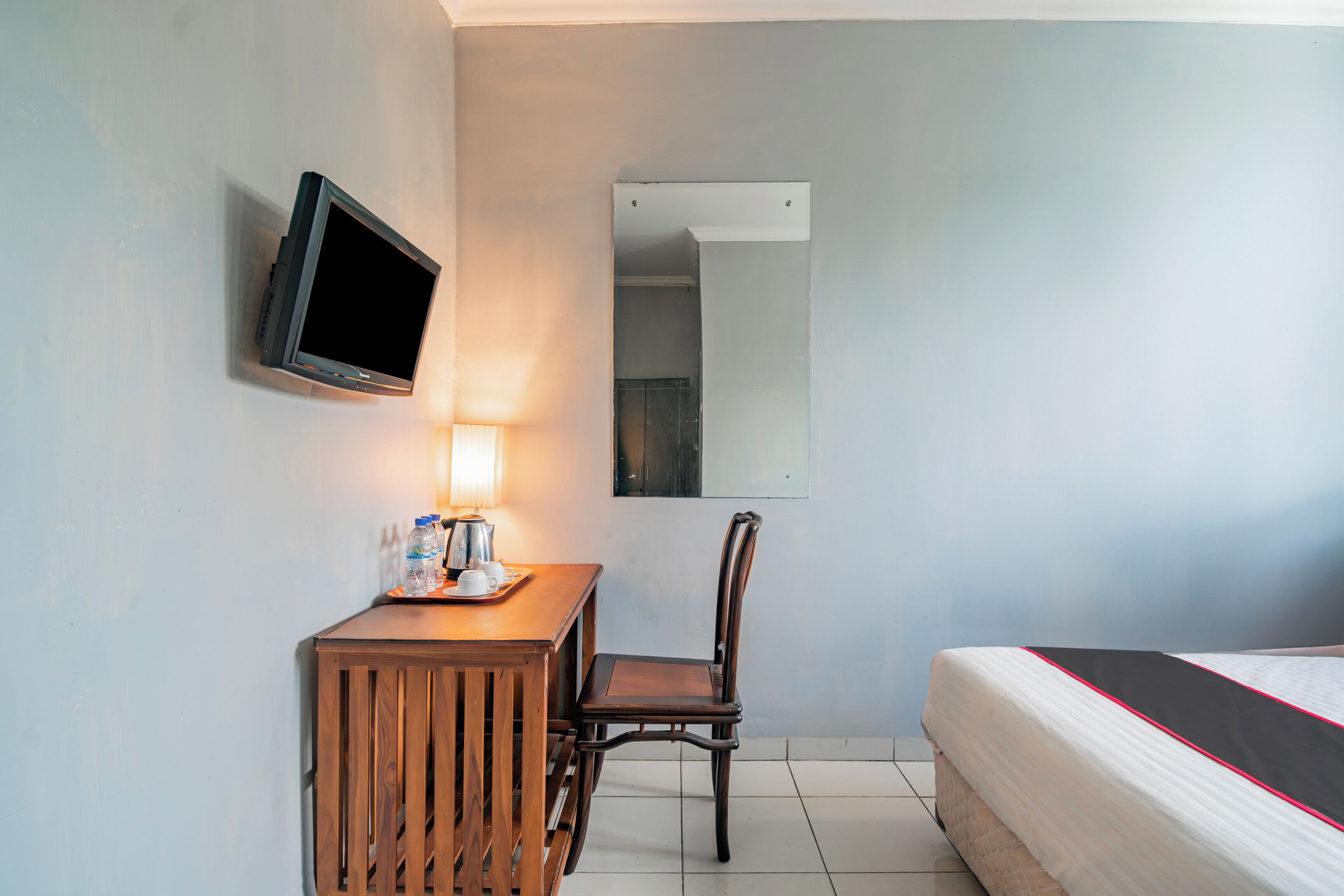 A hotel room with a bed, a desk with a lamp, a chair, a TV mounted on the wall, and a mirror.