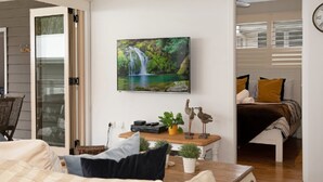 A modern living room with a large TV, a wooden coffee table with decorative items, a bed with a view, and a dining area with a glass door. - Diamond Retreat by HolidayCo. (PEARL BEACH)