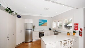 A modern kitchen with white cabinetry, a stainless steel refrigerator, a large island with bar stools, and a window with blinds. - Diamond Retreat by HolidayCo. (PEARL BEACH)
