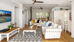A spacious living room with a large TV, a wooden coffee table, striped sofas, and a dining area with a table and chairs. - Diamond Retreat by HolidayCo. (PEARL BEACH)