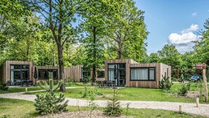 Moderni puinen mökki, jossa on suuret ikkunat, ulkotila istuimineen ja hyvin hoidettu puutarha.