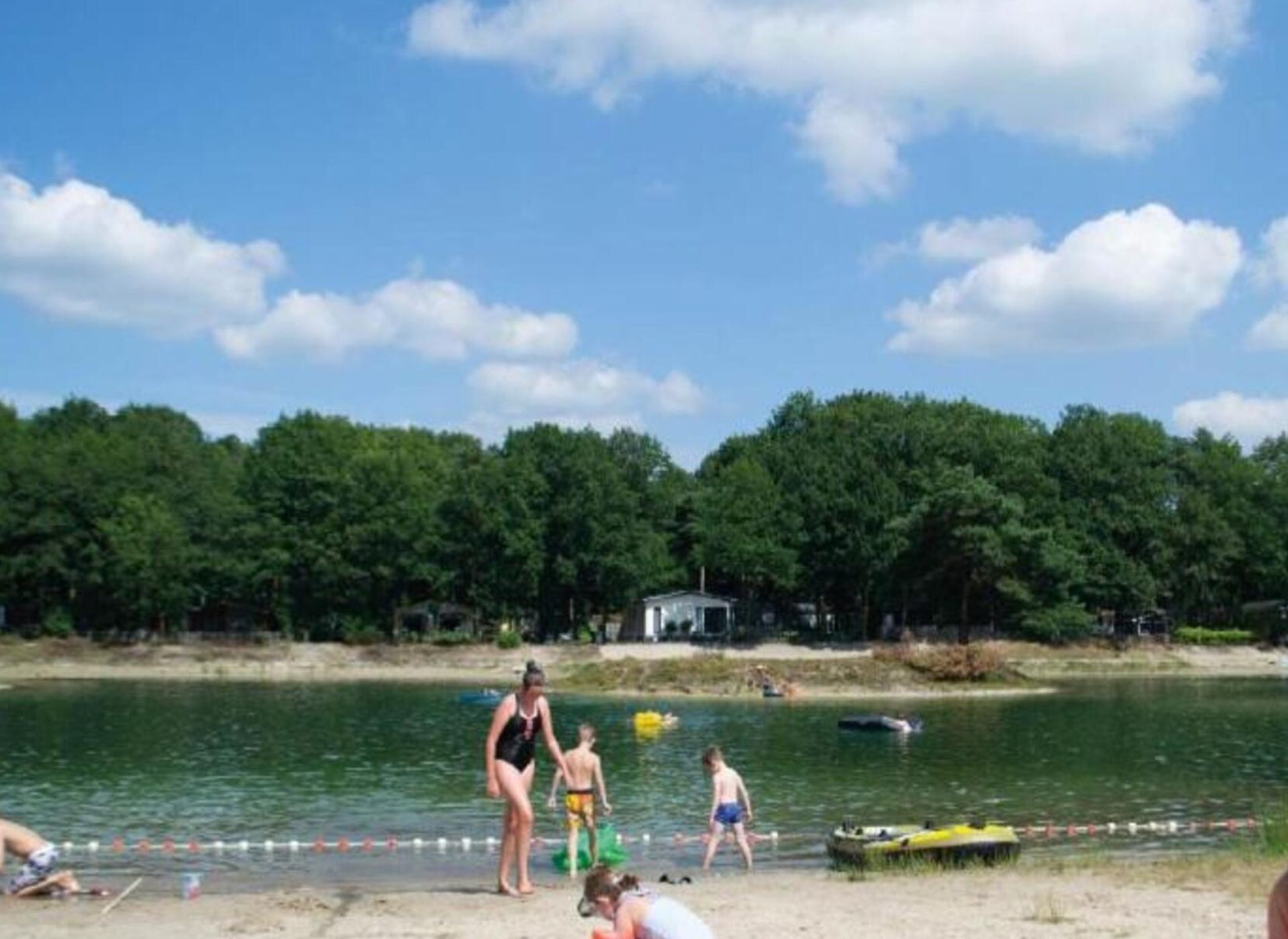 Een strand met mensen die zwemmen en spelen, een steiger met boten, en een bebost gebied op de achtergrond.