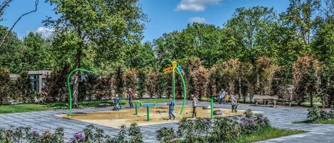 A playground with green structures and a yellow slide, surrounded by trees and a paved area.