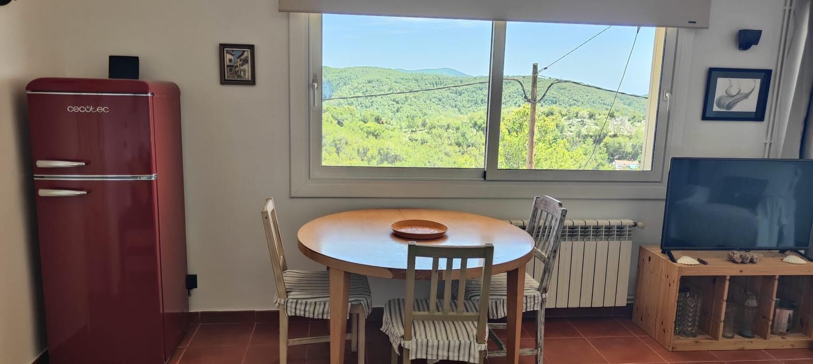 A room with a round wooden table, two striped chairs, a red refrigerator, a television on a wooden stand, and a window with a view of green hills.