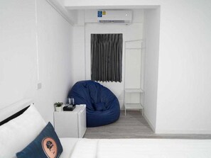 A compact bedroom with a bed, a small bedside table, a blue bean bag chair, a window with dark curtains, and a wall-mounted air conditioner. - RQ North Pattaya By Collection O (Pattaya)