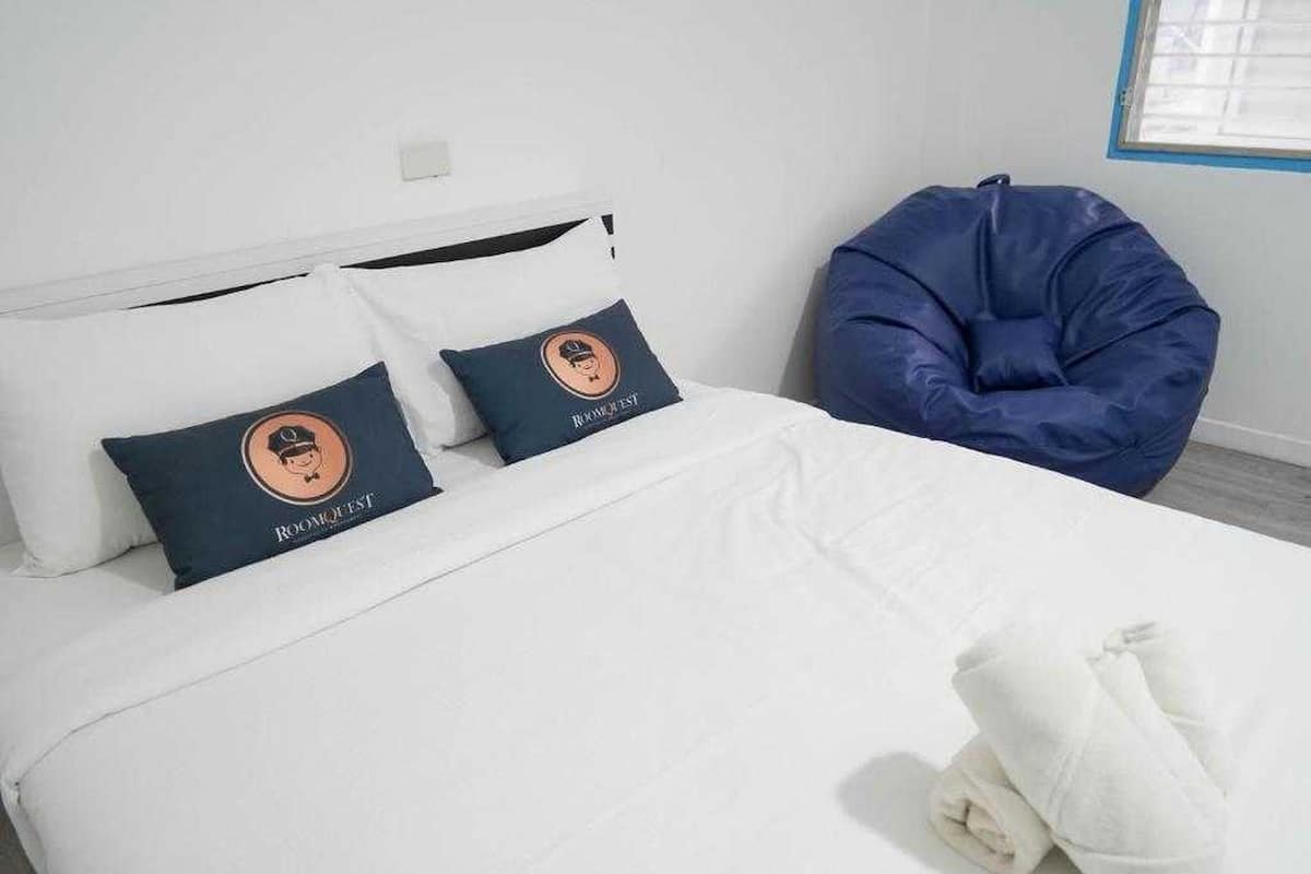A bed with white bedding and two pillows with a logo, a blue bean bag chair, and a window with blinds.