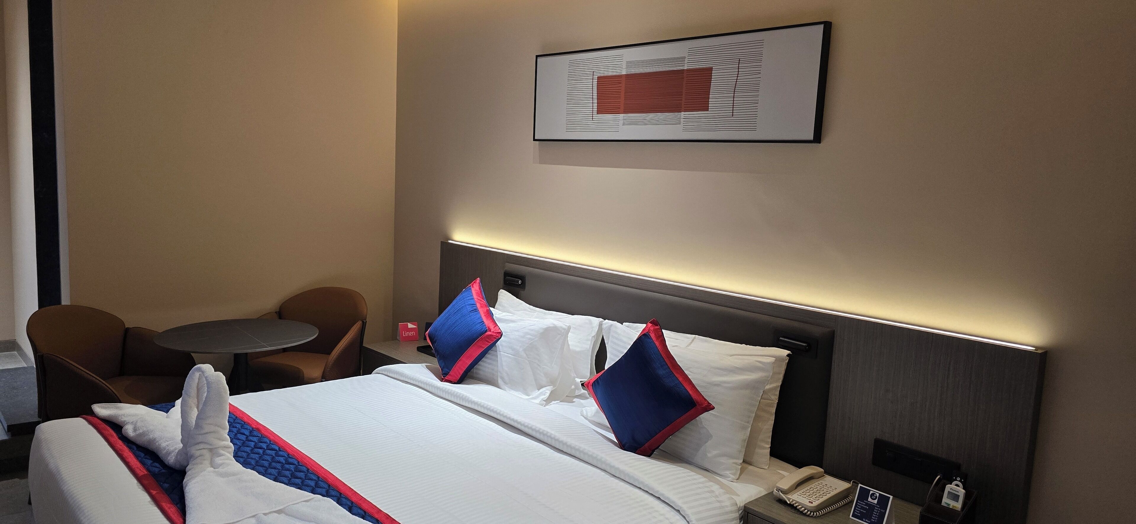 Een hotelkamer met een net opgemaakt bed, een nachtkastje met een telefoon, een klein tafeltje met een stoel, en een ingelijst beeld aan de muur.